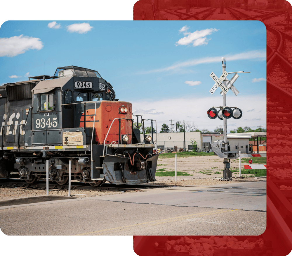 A cropped image showing a black and red diesel locomotive (Engine 9345) approaching a road, with a railroad crossing signal and flashing red lights visible to the right of the engine.